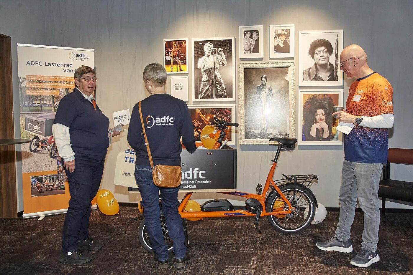 45. ADFC-Bundeshauptversammlung 2025 Drei Personen informieren sich über das orangefarbene ADFC‑Lastenrad. Im Hintergrund sind Roll‑ups zum ADFC‑Lastenrad, Luftballons und mehrere gerahmte Fotos von Musikerinnen und Musikern an der Wand zu sehen.