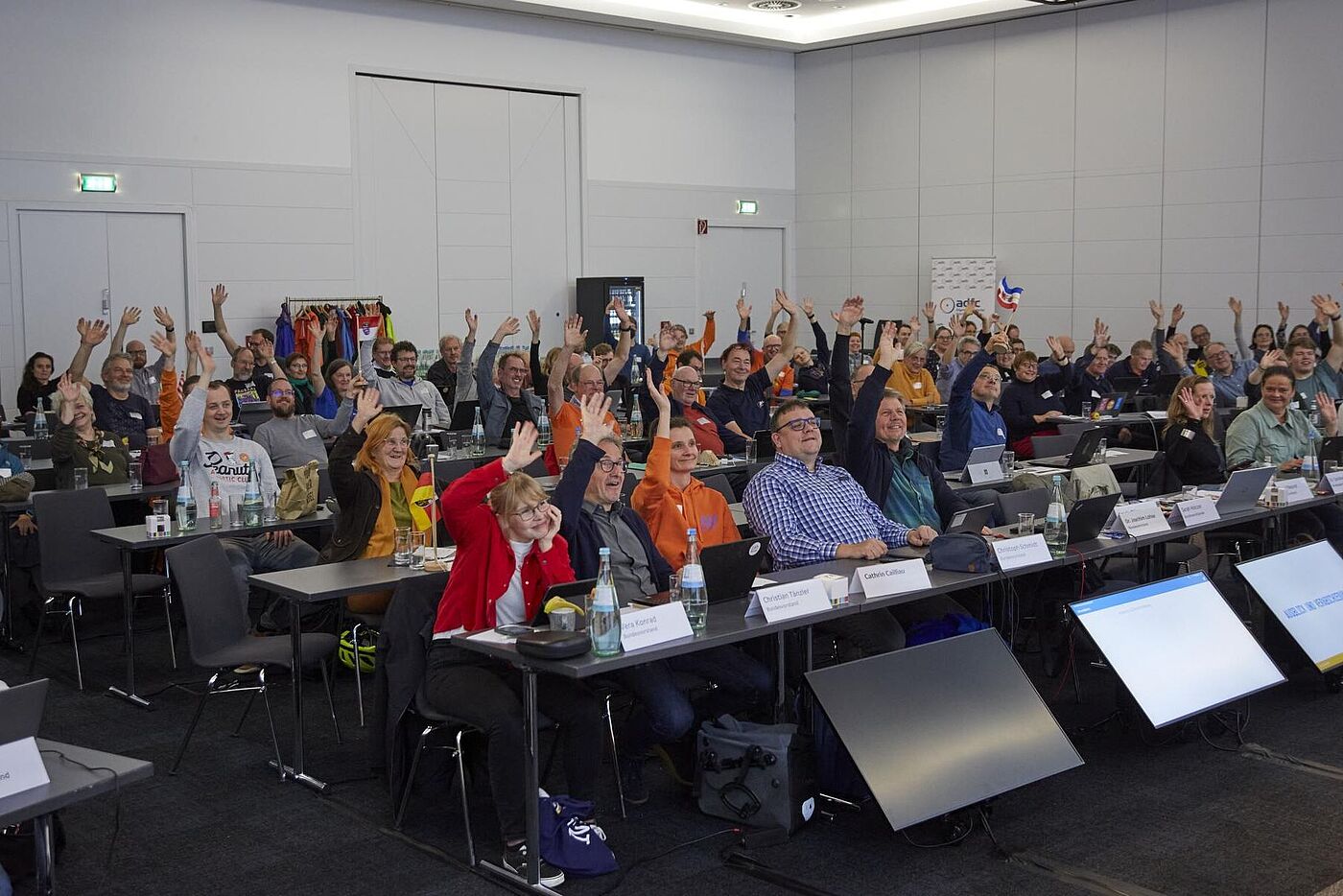 45. ADFC-Bundeshauptversammlung 2025 Die Teilnehmenden sitzen im Saal und heben gemeinsam Hände in die Luft.